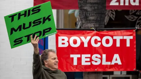 Getty Images A man holds a placard saying "This Musk Stop" in front of a banner reading "Boycott Tesla"