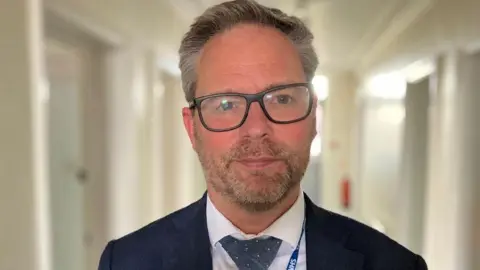 BBC Head and shoulders picture of Martin Hodgson. He has greying hair, a short beard and glasses and is wearing a dark blue suit and tie with a white shirt and NHS lanyard