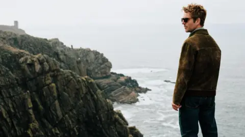 UKTV Actor Damien Molony in character as Jim Bergerac looks over part of Jersey's coastline. He has a brown coat on and dark sunglasses. Rugged rocks are dotted along the coast. It is a gloomy day as waves crash into the rocks.