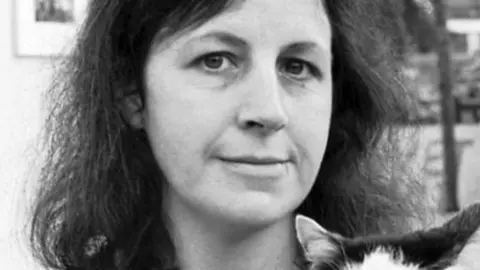 BBC Black-and-white photograph of a woman holding a black-and-white cat. She faces the camera with a calm expression, and the background shows a softly focused indoor gallery or museum space. You can only see the cat's ears.