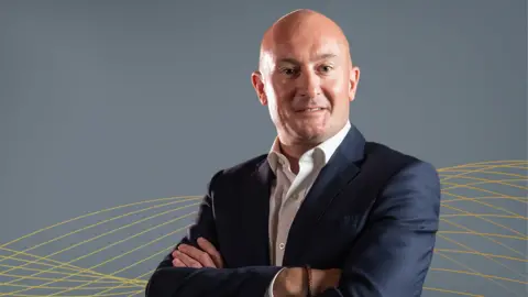 Proserv David Larssen is a bald man in a blue suit and open-necked white shirt. He is standing with his arms folded, turned to his left. The backdrop is a grey wall with yellow patterns.
