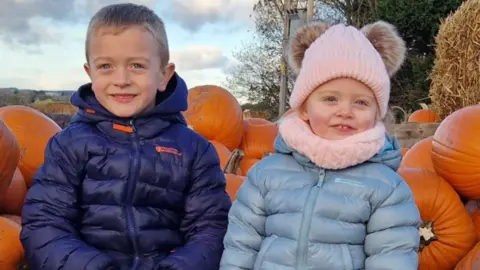 Famile handout Ted and Hollie sit in a pumpkin patch surrounded by pumpkins. Ted wears a dark blue puffer jacket, he has short blonde hair. Hollie wears a light blue puffer jacket, a pink bobble hat and a matching pink scarf. They both smile at the camera 