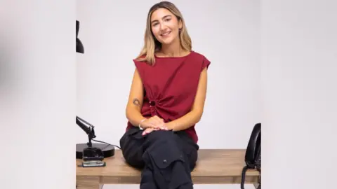 Intotum Hanan smiles for a portrait picture. She has long blonde hair and wears a red sleeveless top. She is in front of a plain white backdrop.