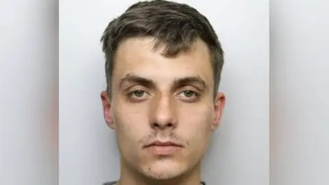 West Yorkshire Police Custody shot of Regan Kemp. He has short brown hair, has a serious expression and is stood in front of a grey background. 