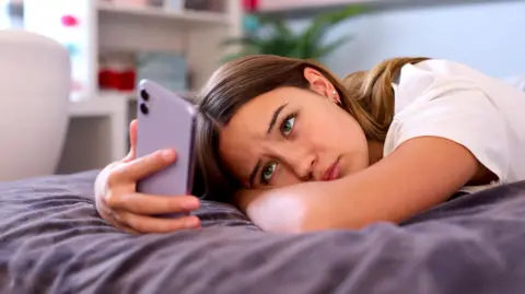 Getty Images A teenage girl is lying on a bed with purple covers looking anxious as she looks at her mobile phone, which is also purple. She is wearing a white top and the room behind her is blurred.