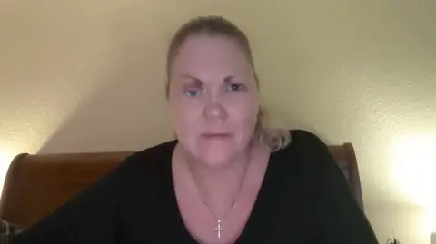 BBC A woman sat on a bed smiling at her computer camera. She is wearing a black long sleeve top and wearing a cross necklace.