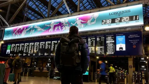 Glasgow 2026 A man with a backpack standing in a railway station, looking up a the large departure boards and the countdown clock of the Commonwealth Games - a blue and purple designed illustration of athletes playing sport, with a clock ticking down to the Games starting. 
