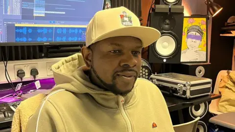 Tom Jackson/BBC Xidus Pain in a recording studio. He is wearing a yellow hoodie with a yellow baseball cap and has a short neat black beard. Behind him is a mixing desk, speakers and a computer screen.
