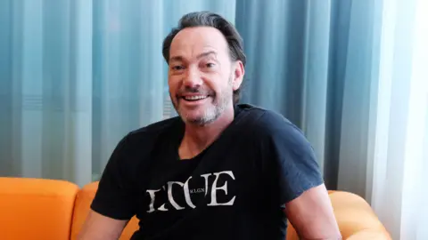 BBC Craig Revel Horwood sat on an orange sofa wearing a black T-Shirt that says "Vogue", smiling at the camera. He has stubbly facial hair and short dark hair with a few grey hairs.