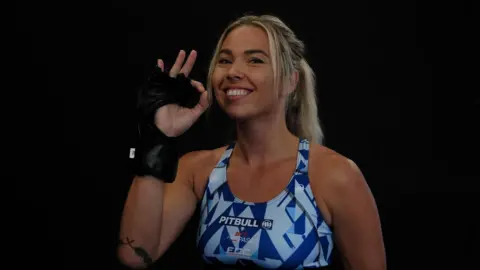 FCC Mia Trevorrow, with her hand making an ok sign. She is wearing a black fingerless glove on her hand. She it wearing a blue sports bra. She is smiling at the camera and her hair is blonde and in a ponytail. The background behind her is black. 