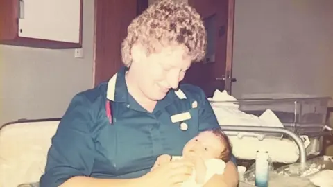 Diane Hele A colour photograph, possibly from the 1980s, of a woman smiling down at a baby she is holding in her arms. She is wearing a dark blue midwife's tunic and has short very curly blonde hair. The baby has dark hair and its eyes are open.