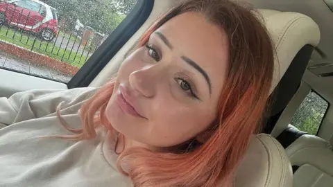 Supplied Lauren smiles slightly in a selfie. She is sat in a car with cream leather seats and it is raining outside. She has red/pink hair with dark roots and make up on. 