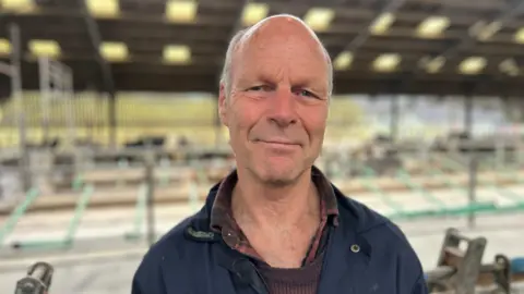 BBC David Finlay wearing a blue boiler suit and burgundy shirt and jumper, smiling inside the cow shed