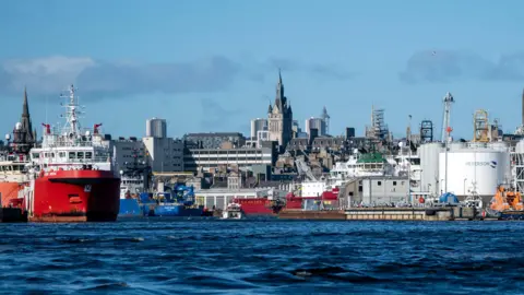 PA Media A view of Aberdeen waterfront and cityscape from close to the water just off shore. We can see two red-hulled ships, grey fuel storage tanks and grey granite buildings behind. It is a sunny day and the sea is very blue.