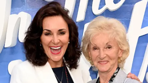 Getty Images Shirley Ballas and her mother smile as they pose together at a photo call at an event. They stand in front of a blue and white backdrop and both wear smart suit jackets.