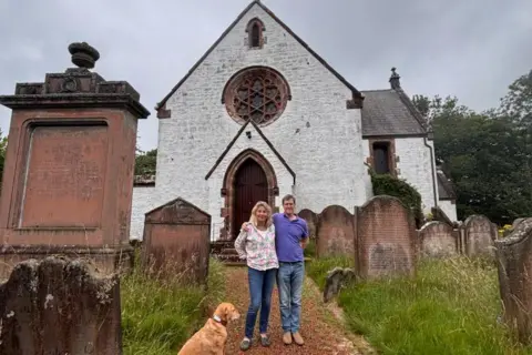 Mitchell family Kirsteen Mitchell and her husband Andrew standing in front of a whitewashed church surrounded by gravestone with a golden-coloured dog by their sides 