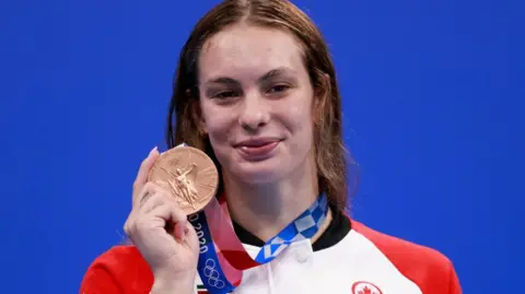 AFP via Getty Images Penny Oleksiak stands against a blue background and smiles as she holds up a bronze medal at the 2020 Tokyo Olympic Games. She is wearing a red and white jacket. 