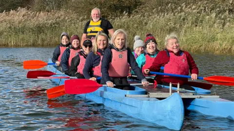BBC Laughing and smiling 10 women sit side by side in a blue bell boat.  Pete Cogley from Paddlers for Life stands at the helm behind them wearing a yellow life vest.  The women are all wearing red life vests and holding red oars. Pete has grey hair and a beard and is waring a black t shirt with a yellow life vest. 