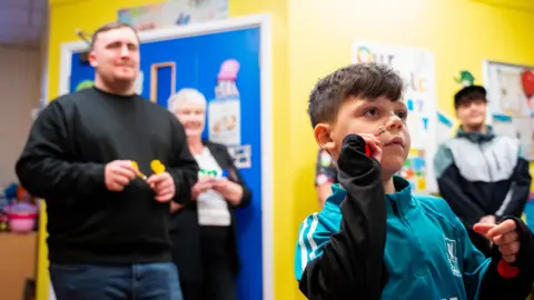 Warrington and Halton Teaching Hospitals Charity In a hospital ward, a dark-haired boy wearing a blue/green tracksuit top is poised to throw a magnetic dart. Behind him is Luke Littler, waiting his turn with more darts. Another adult stands behind smiling in front of a bright blue door in the yellow-walled room. An older boy watches from the other side of the child, and smiles.