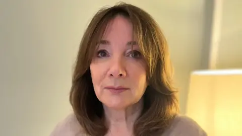 BBC A woman in her 60s, with brown mid-length hair and a beige jumper looks at the camera with a serious expression. She appears to be in a living room, with cream walls and a cream lamp on behind her.