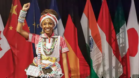 Gallo Images via Getty Images A woman wearing traditional clothing poses with her fist raised. She stands in front of a line of flags from different countries.