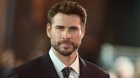Getty Images Liam Hemsworth in black suit and white shirt and black tie with white pattern at the premiere of the show's fourth season smiling at cameras. He has facial hair and short brown hair.