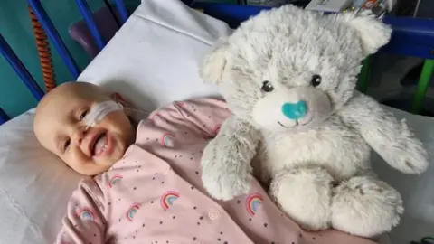 Cancer Support UK Melody, wearing a pink baby grow, lies in a cot smiling broadly. A white teddy bear is beside her.