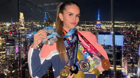Lucy Scott Lucy Scott has eight medals hanging from her neck. They vary from gold, silver and bronze with pink and blue coloured ribbons. She is stood in front of the view of New York City lit up at night. 