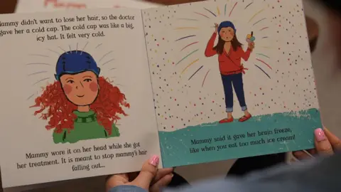 BBC Leanne is reading the children's book. There is an illustration of a woman with curly hair wearing a cold cap. Text around it reads: "Mammy didn't want to lose her hair, so the doctor gave her a cold cap. The cold cap was like a big, icy hat. It felt very cold. Mammy wore it on her head while she got her treatment. It is meant to stop mammy's hair falling out... Mammy said it gave her brain freeze like when you eat too much ice cream!"