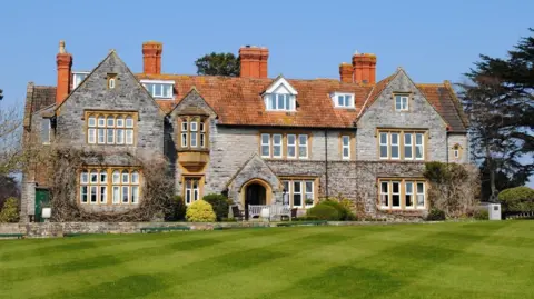 Millfield School An exterior view of Millfield School, showing an old stone building in the sunshine, with blue skies behind it and large trees to each side. In front is an immaculately mown lawn cut with a diamond pattern.