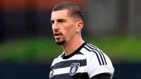 SNS A footballer with short dark hair and a beard and moustache. He is standing on the pitch wearing a black and white hooped kit.