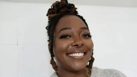 John Fairhall/BBC Shea Osei smiles at the camera. She has black hair in twists and a ring in her nose. She is wearing a grey sweater and is standing in front of a white wall.
