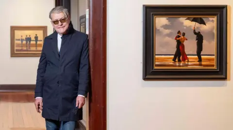 PA Media The late Jack Vettriano in a navy coat, leans on a doorway beside a white wall bearing his best-known painting The Singing Butler