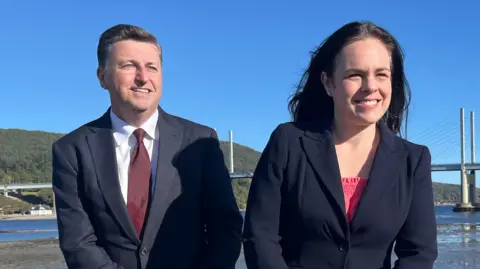BBC Douglas Alexander, wearing a navy suit and burgundy tie, sitting on a wall next to Kate Forbes who is wearing a dark blazer and pink top. Behind them is a bridge and water.