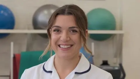 Day One Trauma Chloe Boobier dressed in her white physio uniform and is smiling for the camera. She has long brown hair that has been tied back.