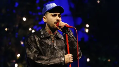 Getty Images Zayn Malik wears a leather jacket and a blue hat. He is singing into a red microphone on a red microphone stand. 