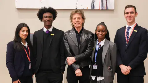 Pete Bresser Sir Mick Jagger wearing a dark shirt and leather jacket. Four older children wearing school uniform and lanyards are stood either side of him. All are smiling at the camera.