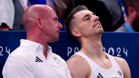PA Media Scott Hann and Max Whitlock sitting next to each other in an arena at the 2024 Paris games. Mr Hann is wearing a white Team GB polo shirt and talking in the ear of Whitlock, who is wearing a white vest. He looks upset and is leaning back.