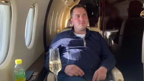BBC/SpunGold TV Miles Hart, a young man with short brown hair, wearing a quarter-zip blue top, sitting in a cream leather seat on a small, luxurious jet, with a glass of what appears to be champagne in front of him.