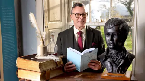 Colin Hattersley A man in glasses with a red tie and a brown jacket and cream shirt opens a book next to a bust of Robert Burns and some books and quill pens