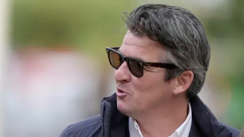 PA Media Joey Barton smiles as he speaks to someone over his shoulder on his way into court. He has shirt dark hair and wears dark shades.
