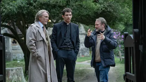 John Wilson/Netflix (Left to right) Daniel Craig and Josh O'Connor and writer-director Rian Johnson are standing just beyond the entrance to a church graveyard with the church itself in the background. Johnson, on the right, is gesturing to Craig and O'Connor, who are listening intently. Craig is wearing a long beige raincoat with shirt and tie, while O'Connor is dressed as a clergyman in dog collar and black shirt, jacket and trousers. 