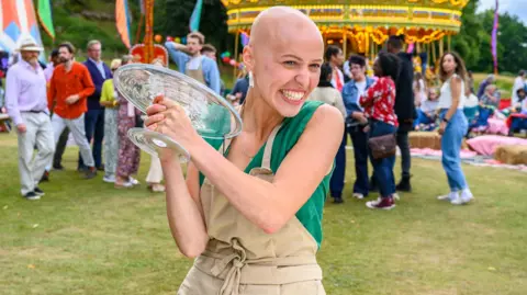 Channel 4 A woman wearing a beige apron and green top is holding two large glass plates outdoors at a lively fairground. In the background, there are colourful tents, a carousel, and groups of people enjoying the event.