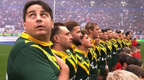 BBC Man in green and yellow Australia rugby strip looks up over his shoulder at the big screen while at the end of the team's line-up for the national anthem.