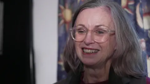 BBC Carolyn Hargreaves, a woman with shoulder-length grey hair wearing glasses and a dark vest over a maroon top, standing indoors against a colorful patterned backdrop featuring abstract sun-like designs.
