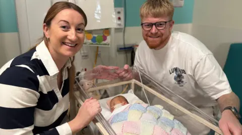BBC A woman and man standing either side of their newborn baby who is being treated in a hospital's neo-natal unit. The baby is asleep under a multi-coloured blanket. 
