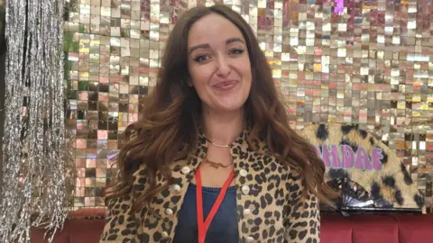 BBC Laura Mears-Reynolds is sitting on a red sofa in front of a mirrored wall. She is wearing a leopard-print jacket and has long, brown hair. She is smiling at the camera.