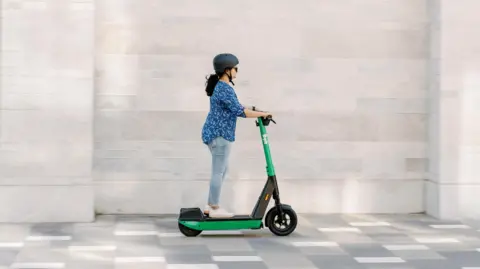 Bolt A woman with black hair and wearing a blue blouse and jeans rides on a green electric scooter with the Bolt logo on the frame. 