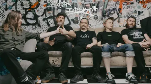 Ellen Dixon Pigs x7 which is made up of five men, all sitting on a sofa in front of a graffitied wall. They are all wearing dark colours and the two on the left are holding their thumbs together like they're in the middle of a thumb war. The man on the far left has long, brown hair and is looking to the side. Next to him, the man has a grey beard and is wearing a black cap. The man in the middle has dark hair which looks to be tied up and a dark moustache. He is wearing a black Korn band t-shirt. The man middle right has light hair and is wearing a grey cap and is looking to the right with his hands in his lap. He is wearing turned up jeans that are exposing the bottoms of his legs. The band member on the far right has chin-length dark hair and a greying beard and is looking into the camera with his hands clasped on his lap. 