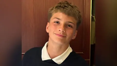 British Transport Police Joshua Travis, a 14-year-old boy with mousey brown hair. He is wearing a school uniform and appears to be standing in front of a wardrobe and he his smiling.